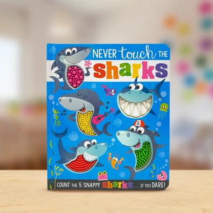 A children's board book titled 'NEVER touch the sharks' standing upright on a light wooden surface. The cover features colourful cartoon sharks, bubbles, and a white shark mouth with teeth on the front.