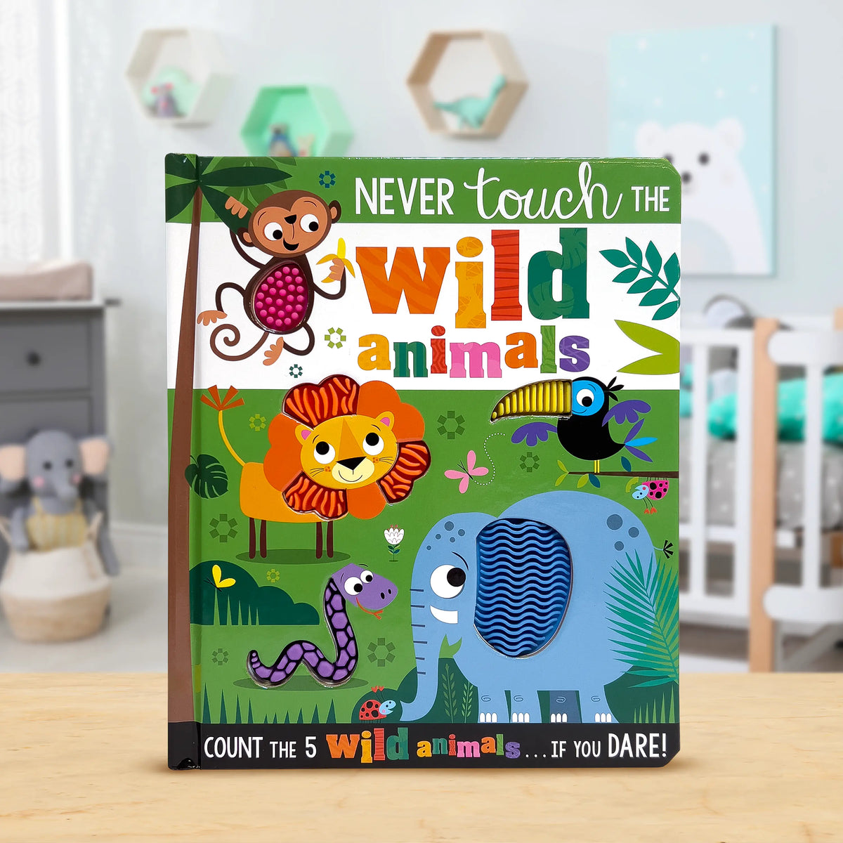 A children's board book titled 'Never touch the wild animals' stands upright on a light wood surface in a softly blurred nursery setting. The cover features colourful cartoon animals including a monkey, lion, toucan, bird, snake, and elephant on a green jungle-style background.