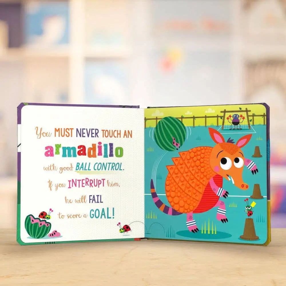 An open children's board book showing a colourful illustration of an armadillo on one page and a rule about ball control on the other, photographed on a light wooden surface with a blurred indoor background.