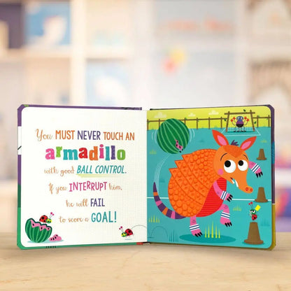 An open children's board book showing a colourful illustration of an armadillo on one page and a rule about ball control on the other, photographed on a light wooden surface with a blurred indoor background.