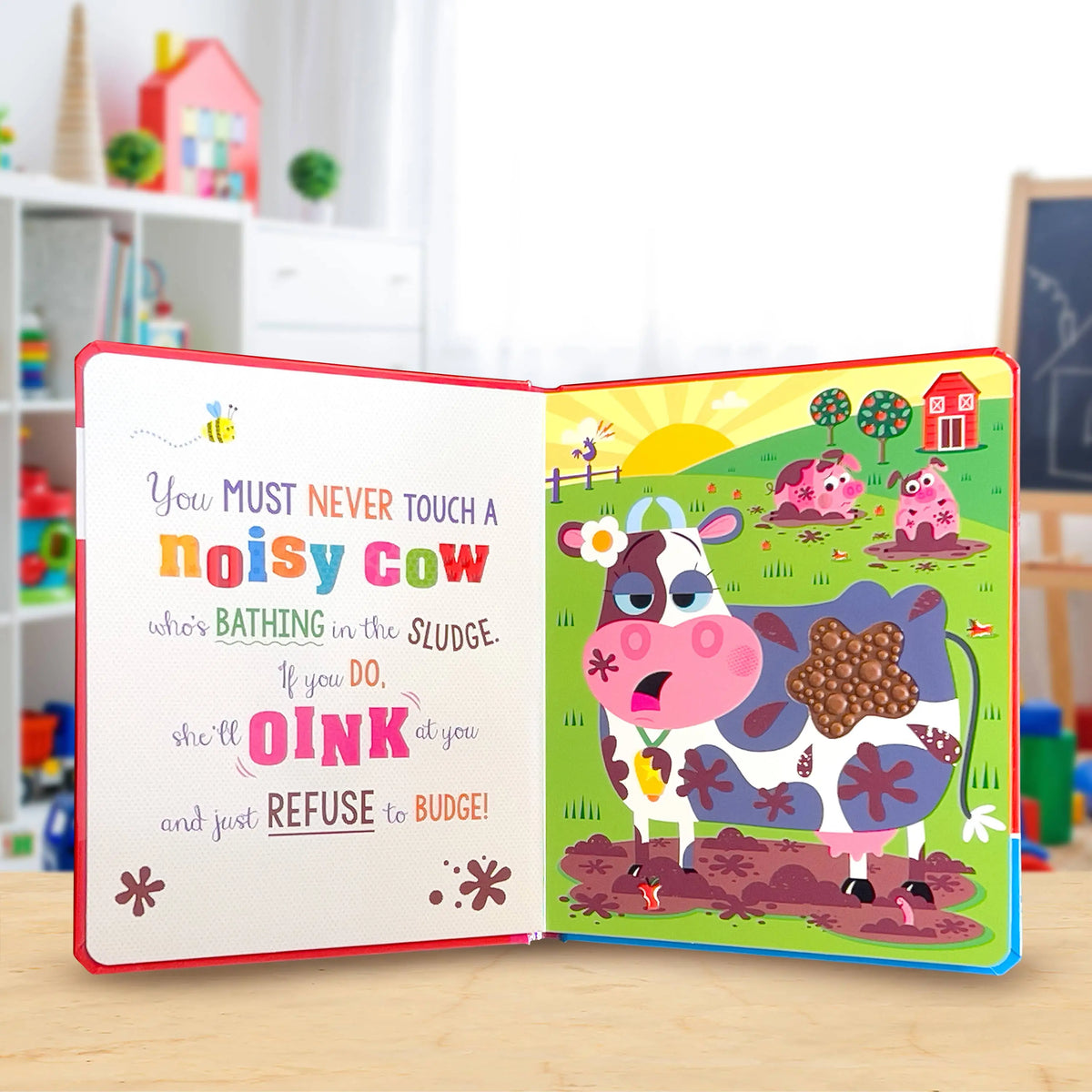 An open children's board book with a colourful pop-up cow scene on the right page and a text-based left page, displayed on a light wood surface with a blurred playroom background.