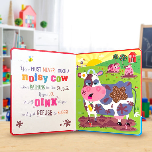 An open children's board book with a colourful pop-up cow scene on the right page and a text-based left page, displayed on a light wood surface with a blurred playroom background.