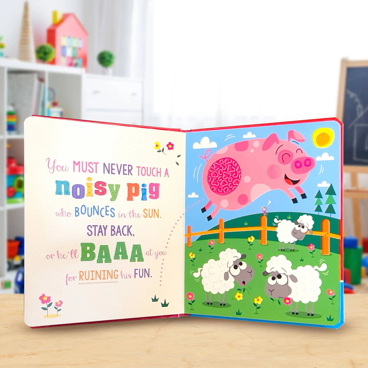 An open children's book on a light wood surface, showing a colourful illustrated scene with a pink pig jumping over sheep in a field and a playful text quote on the left page. The background is a softly blurred indoor room with shelves and toys.