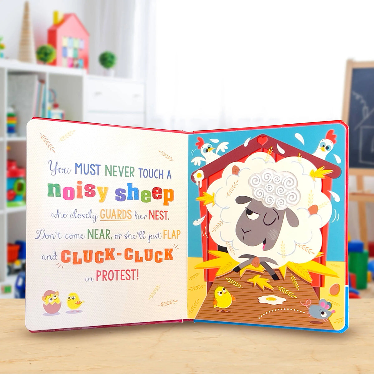 An open children's board book with colourful illustrations and text, showing a sheep in a barn with chicks and a mouse. The book is photographed on a light wooden surface with a blurred playroom background.