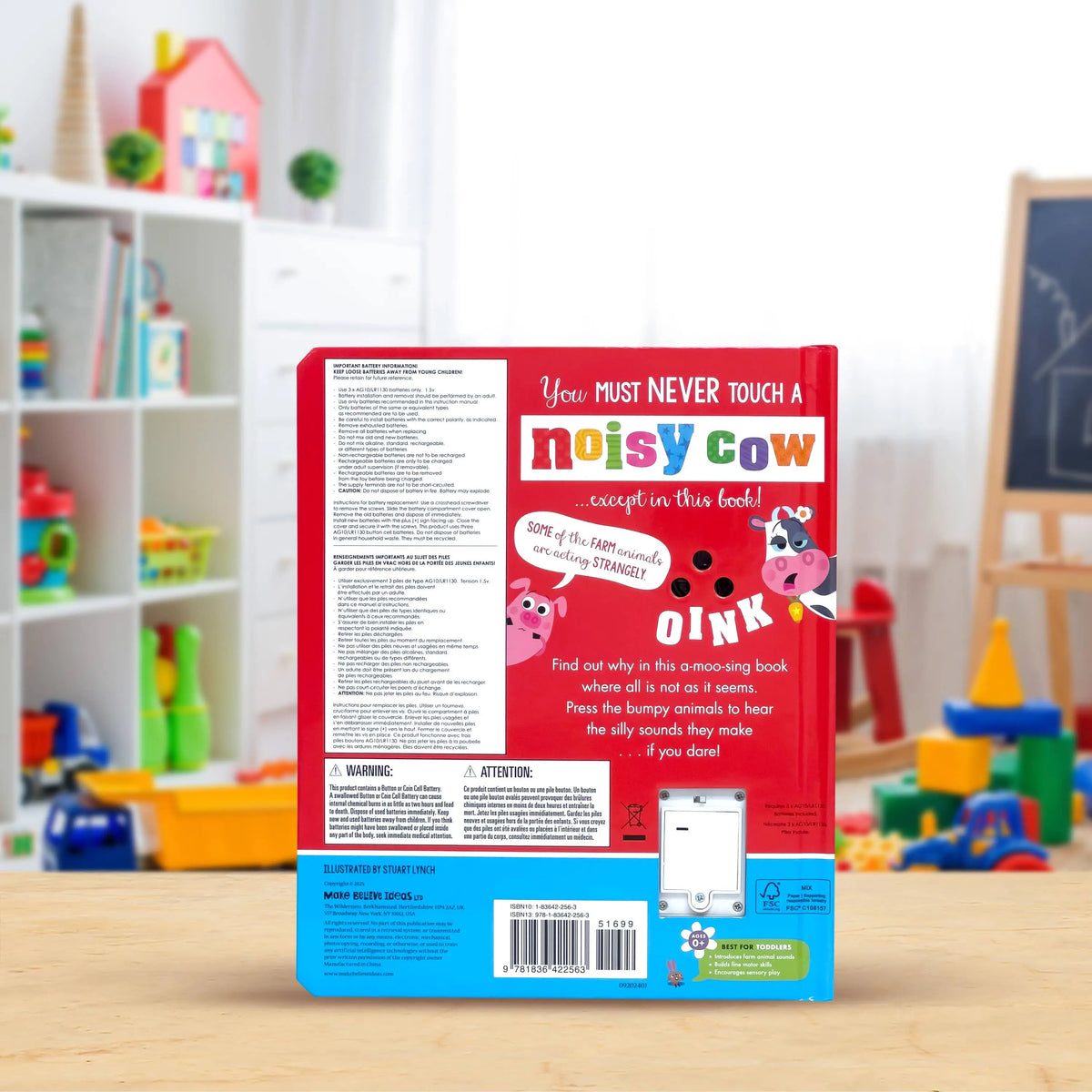 The back cover of a children's book titled 'Noisy Cow' is shown standing upright on a light wood floor in a brightly lit, blurred playroom setting. The cover features a red top half with white text and cow illustrations, a white middle section with safety instructions, and a blue bottom section with a barcode and small product icons.