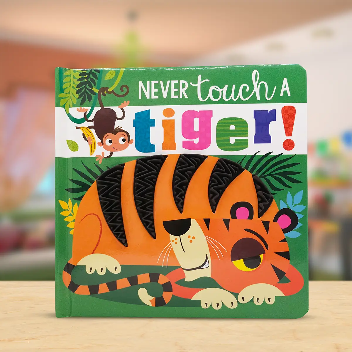 A children's hardboard book titled 'Never touch a tiger!' with a colourful illustrated cover showing a tiger peeking out from behind green leaves, photographed standing on a light wood surface with a blurred indoor background.