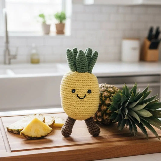A crocheted pineapple toy with a smiling face stands on a wooden cutting board in a bright kitchen setting, surrounded by real pineapples and pineapple slices.