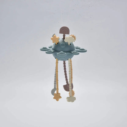 A baby teether with a dusty blue UFO-shaped centre, hanging wooden and plastic dangling elements, and star and cloud shapes, photographed against a plain light grey background.