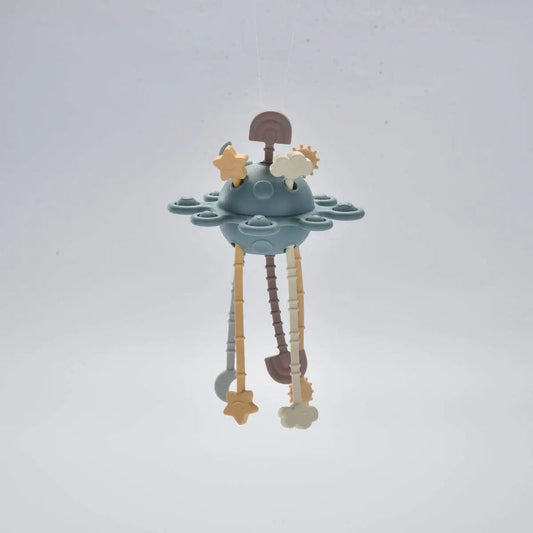 A baby teether with a dusty blue UFO-shaped centre, hanging wooden and plastic dangling elements, and star and cloud shapes, photographed against a plain light grey background.