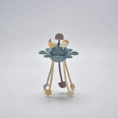 A pastel-coloured baby activity toy shaped like a small space station or UFO, featuring a blue-grey central dome, three hanging legs in beige, brown, and grey, and small star and gear accents.