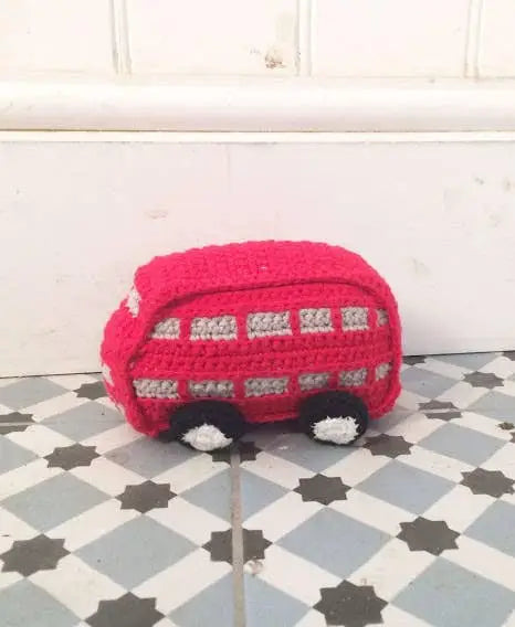 A crocheted red toy bus with white and black details, placed on a gray and white checkered floor against a white paneled wall.