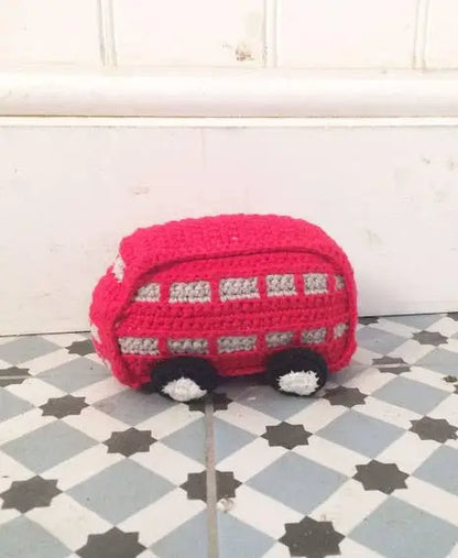 A crocheted red toy bus with white and black details, placed on a gray and white checkered floor against a white paneled wall.