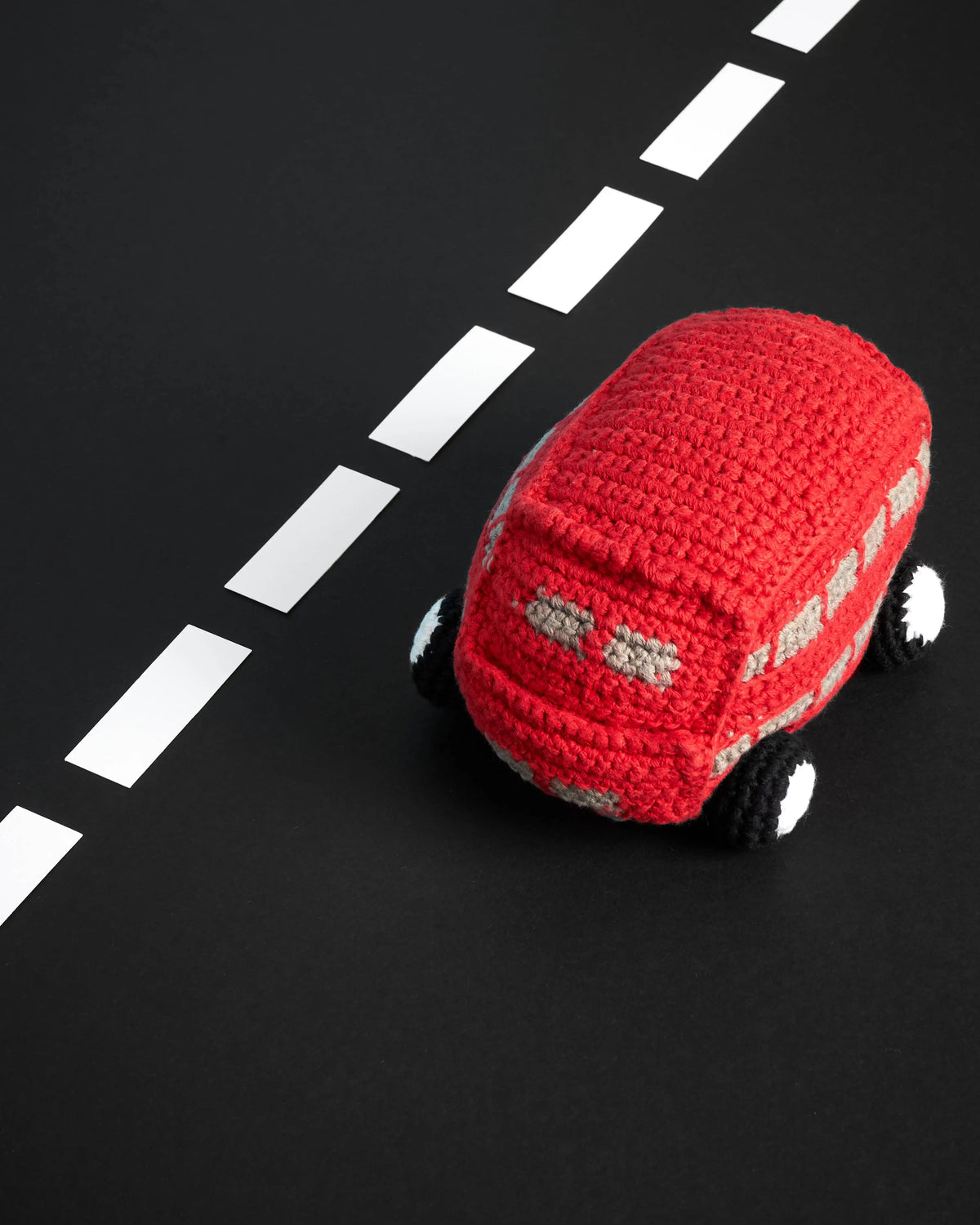 A red crocheted toy car with black and white wheels, placed on a black surface with a white dashed road line.