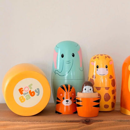 A set of colourful baby nesting dolls and a matching storage container on a wooden shelf against a plain light wall. The set includes a mint green elephant, a yellow giraffe, an orange tiger, a small brown monkey doll, and a round yellow container labeled 'For the Baby'.