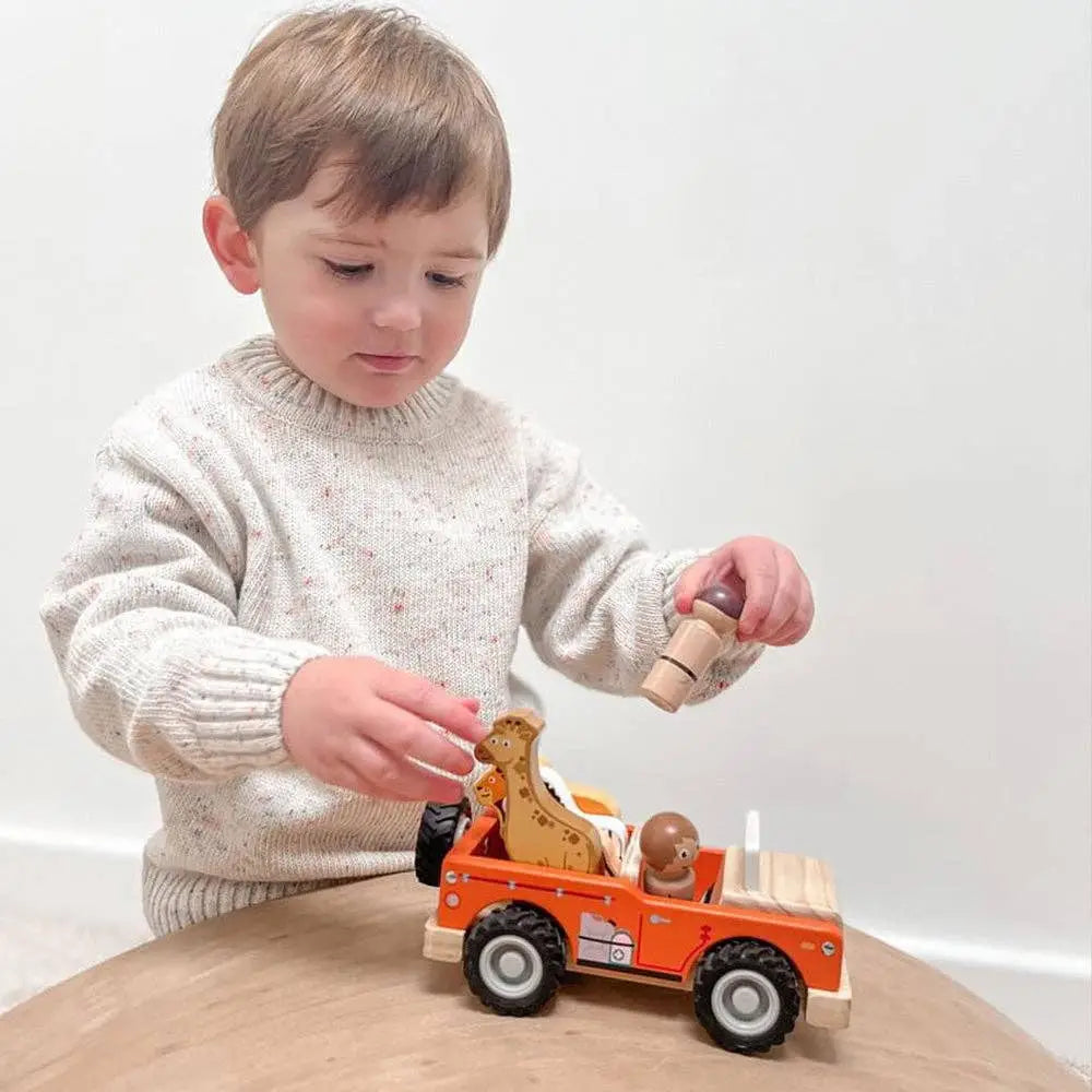 A young child wearing a cream speckled knit sweater is playing with a wooden toy construction vehicle on a light wood table against a white wall.