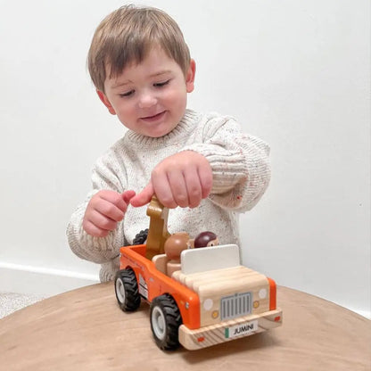 A young child wearing a cream knit sweater is sitting on a wooden table, playing with a wooden toy jeep. The toy jeep is orange and natural wood, with small animal figures inside.