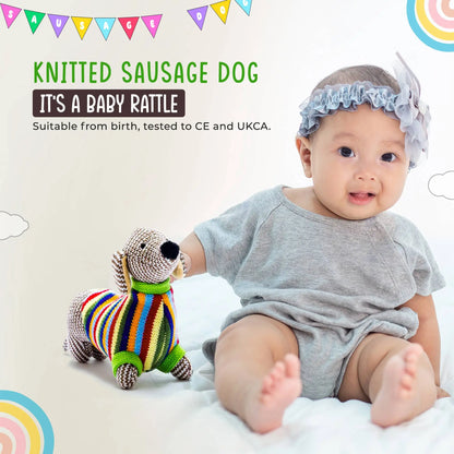 A baby wearing a light blue ruffled bonnet and a grey short-sleeve romper sits on a white blanket next to a knitted sausage dog toy. Text on the image reads 'KNITTED SAUSAGE DOG' and 'IT'S A BABY RATTLE'.