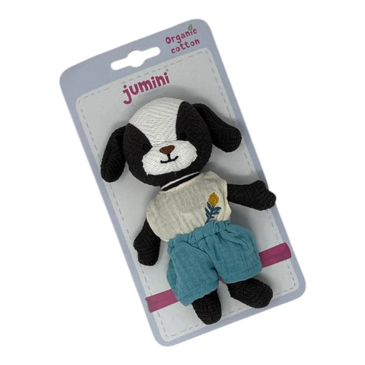 A plush black and white puppy toy dressed in a cream shirt with a small yellow flower and blue shorts, packaged on a light blue card labelled 'jumini' and 'Organic cotton'.