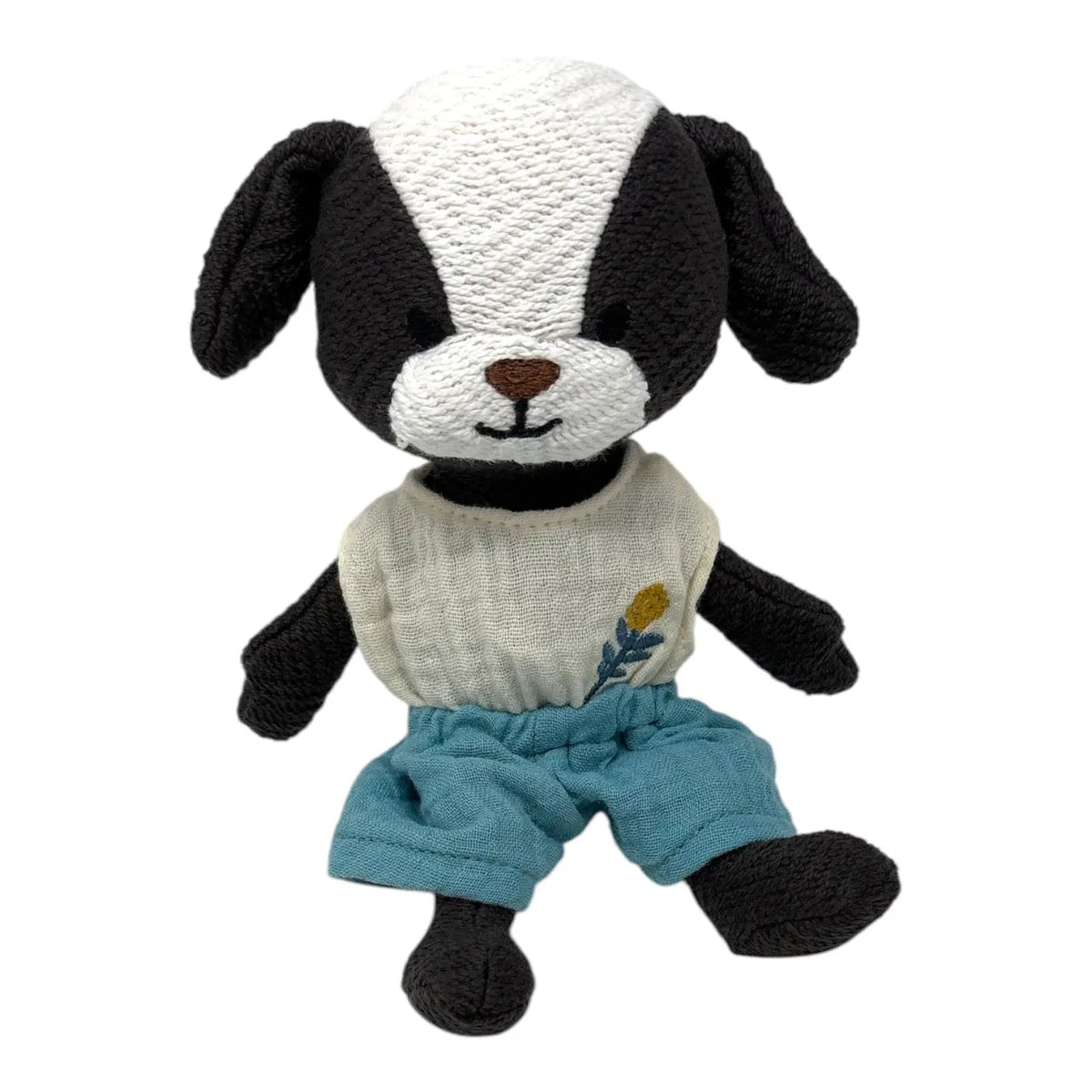 A handmade crocheted plush puppy toy with a black and white face, wearing a cream-coloured shirt with a small yellow flower and blue pants, set against a plain white background.