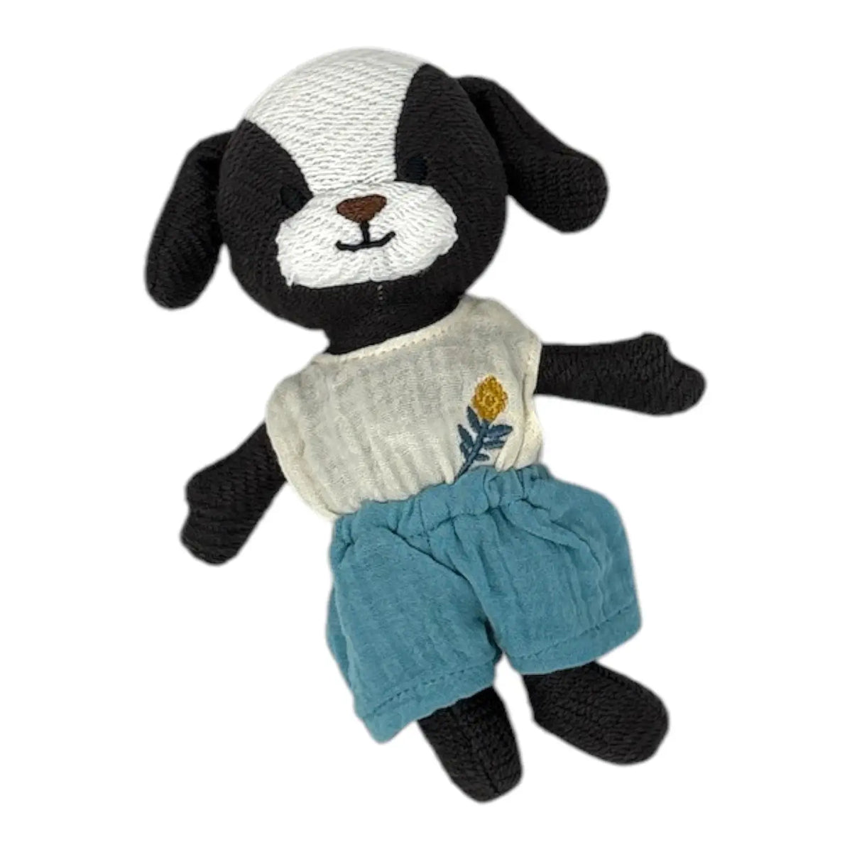 A handmade black and white knitted dog toy wearing a cream-coloured top with a small yellow and blue flower embroidery and teal-blue shorts, isolated on a white background.