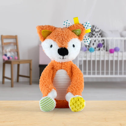 A plush toy fox sitting upright on a light wooden table in a softly blurred nursery setting. The toy has orange fur, a white face and belly, black embroidered eyes and nose, green inner ears, striped green-and-white socks, and a yellow-and-white polka dot bow on its head.