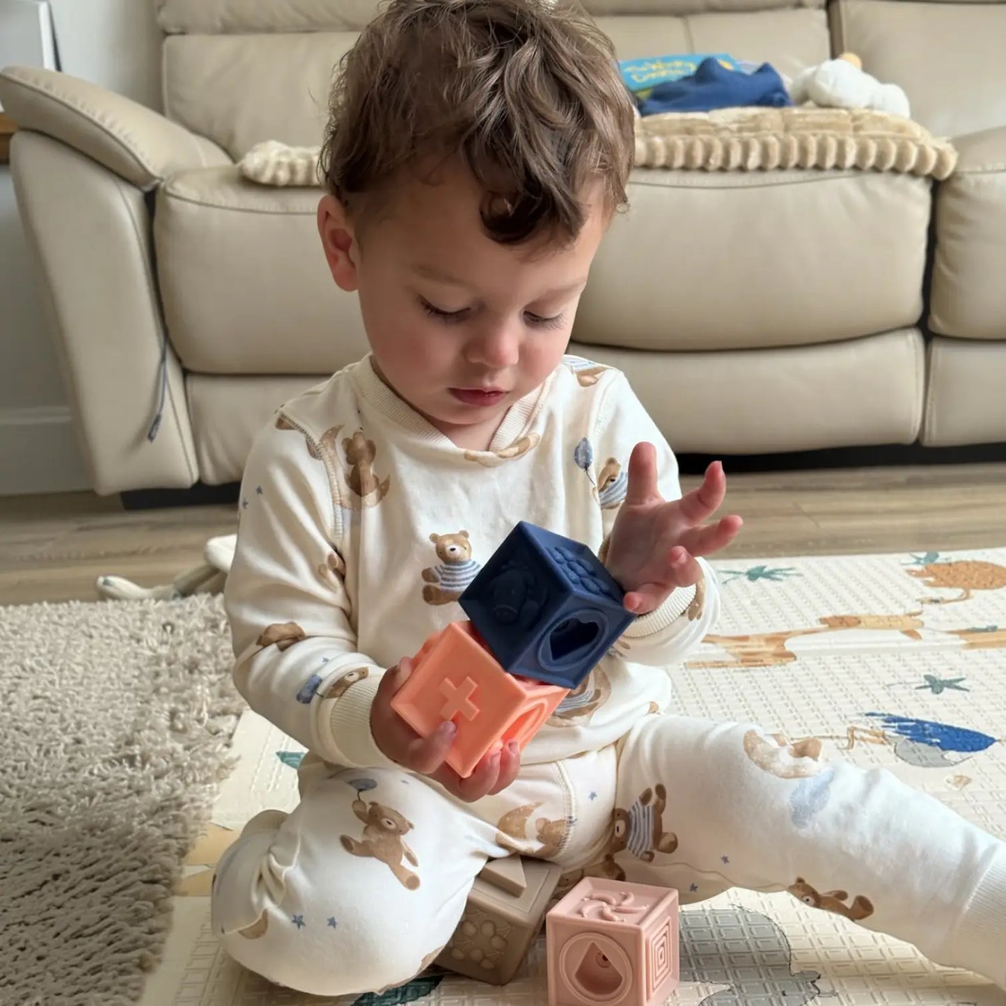 A toddler sitting on a play mat in a living room, wearing a cream pyjama set with teddy bear prints and holding colourful silicone shape blocks.