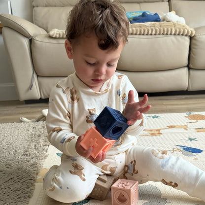 A toddler sitting on a play mat in a living room, wearing a cream pyjama set with teddy bear prints and holding colourful silicone shape blocks.