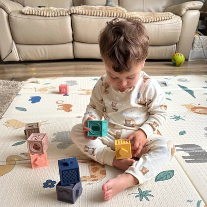 A toddler sits on a patterned play mat in a living room, holding colourful geometric blocks. The child is wearing a cream pyjama set with a brown animal print.