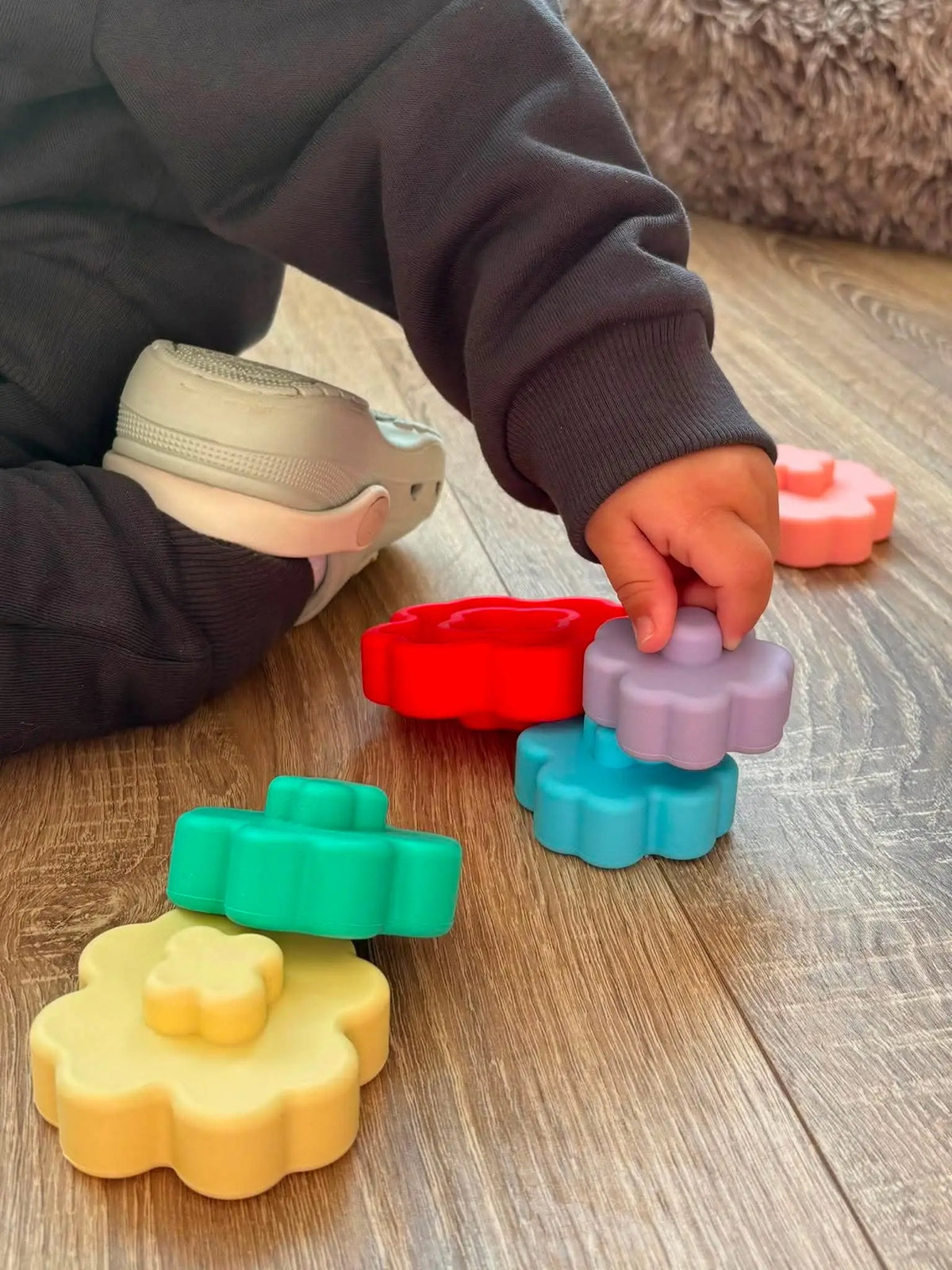 A child wearing a dark sweatshirt and light-coloured shoes is sitting on a wooden floor, stacking colourful plastic flower-shaped toys. The toys include yellow, green, red, blue, purple, and pink pieces.