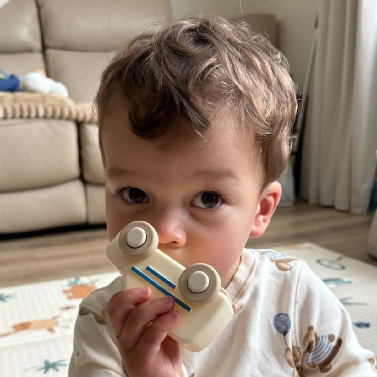 A toddler boy holding a small cream-coloured toy ambulance with beige wheels and blue stripes, posing in a cozy living room setting.