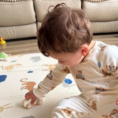 A baby wearing cream-coloured pyjamas with a brown teddy bear pattern sits on a play mat, holding a small  toy car in a living room setting.
