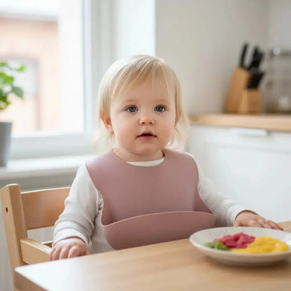 Child sitting at a table with a plate of food, wearing a pink bib.