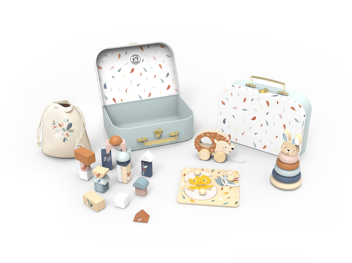 A children's play set arranged on a clean white background, including two illustrated metal cases, a drawstring bag, wooden house blocks, a small plush toy, a stacker ring toy, and a rectangular activity mat.