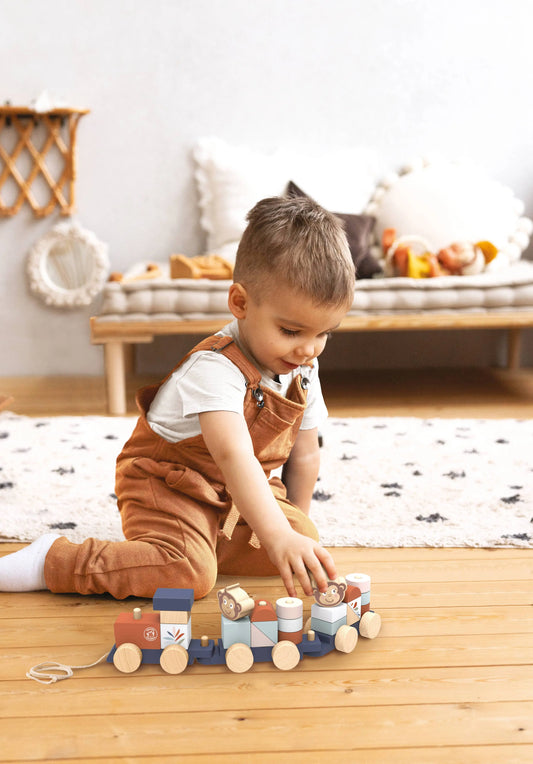 A toddler wearing brown corduroy overalls sits on a wooden floor, playing with a colourful wooden toy train in a bright, modern room.