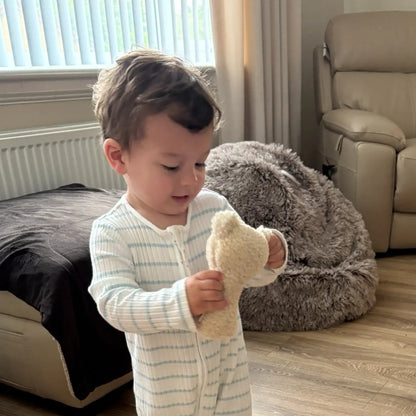 A toddler wearing a white and light blue striped long-sleeve footless onesie stands indoors holding a small beige plush toy. The room has wood flooring, a beige recliner, a gray bean bag chair, and a window with blinds in the background.