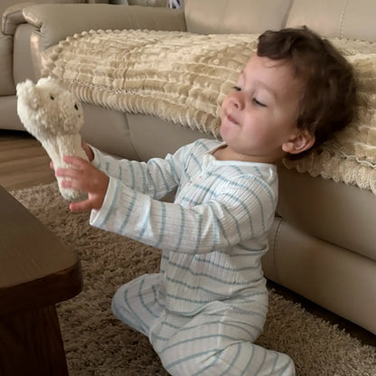 A toddler wearing a light blue and white striped long-sleeve footed pajama sits on a beige carpet in a living room, holding a small beige plush toy near a beige sofa.