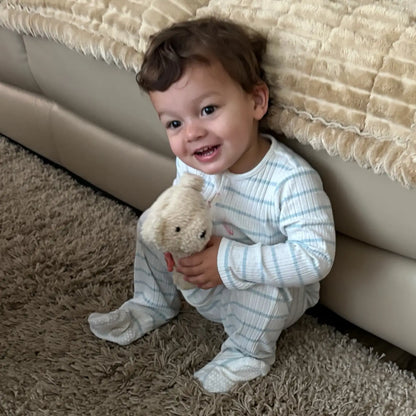 A smiling toddler sitting on a shag rug in front of a beige sofa, wearing a light blue and white striped long-sleeve footed pajama and holding a small plush toy.