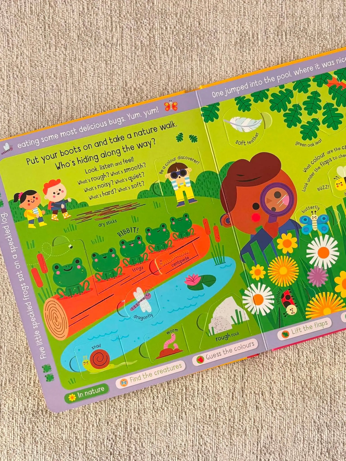 A children's activity book spread photographed on a beige fabric surface. The pages show colourful illustrated scenes with text prompts, including a nature walk, pond creatures, flowers, and a character in a treehouse.