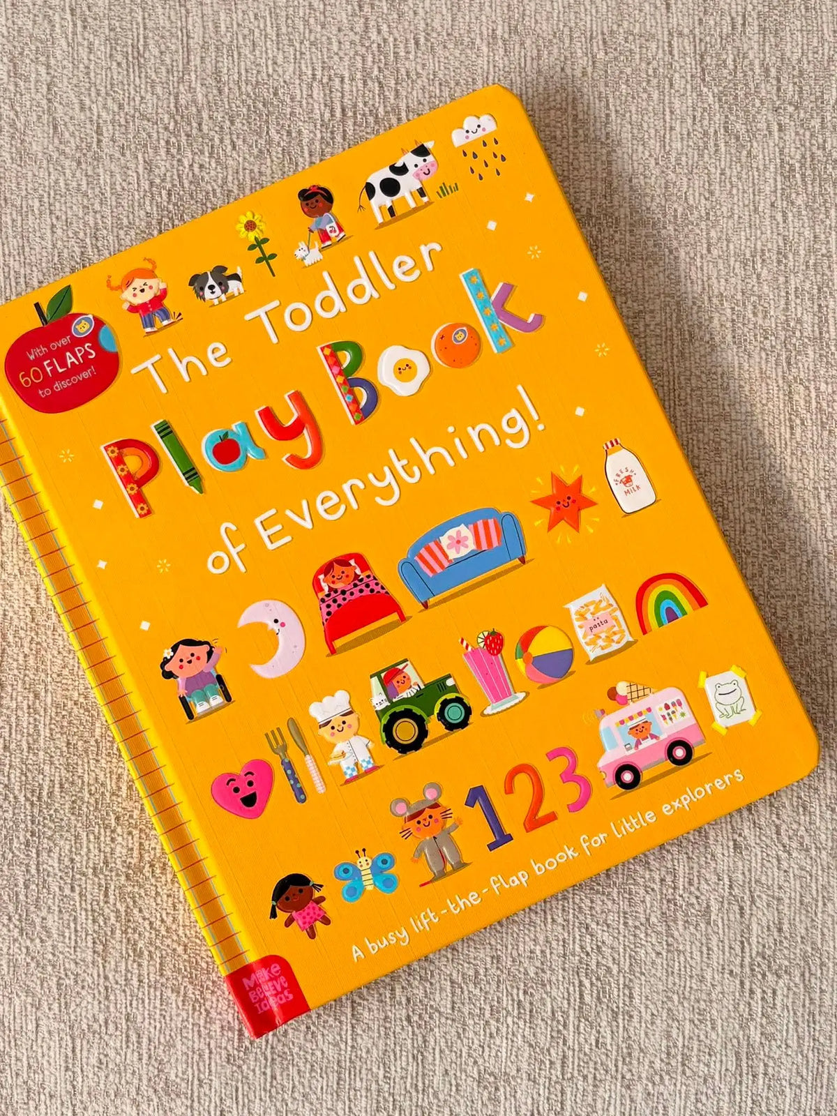 The Toddler Play Book of Everything! in a bright yellow hardcover, featuring colourful illustrations of children, animals, vehicles, and numbers, photographed on a textured beige fabric background.