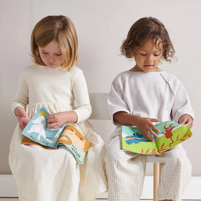 Two young children sitting on chairs and reading colourful picture books in a bright, neutral room. The child on the left wears a cream long-sleeve dress and holds a blue and orange book.