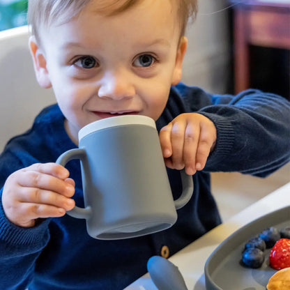 A toddler holding a matte grey silicone mug with a white lid, wearing a navy blue sweater in a bright indoor setting.
