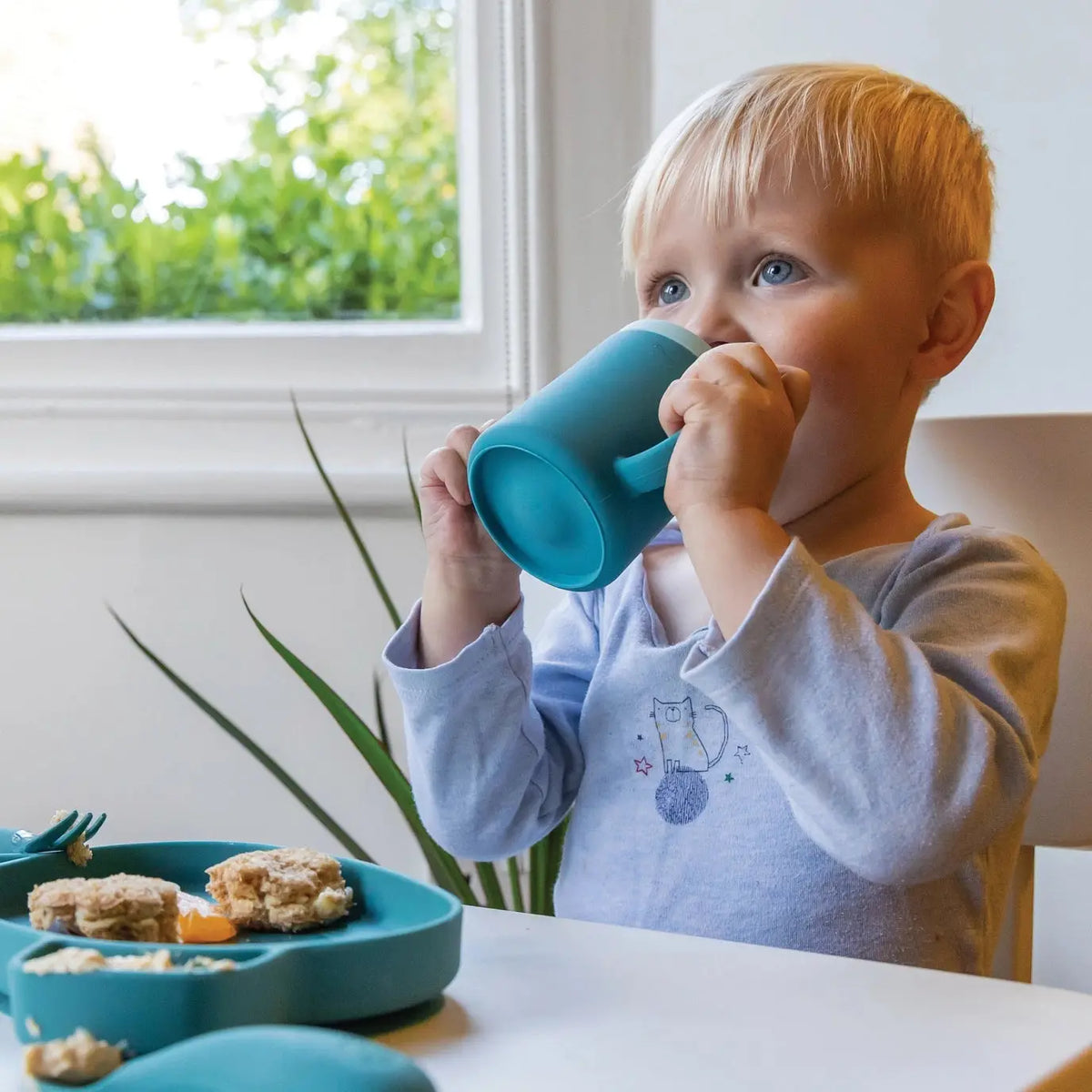 Child drinking from a blue cup at a table with a window and plant in the background