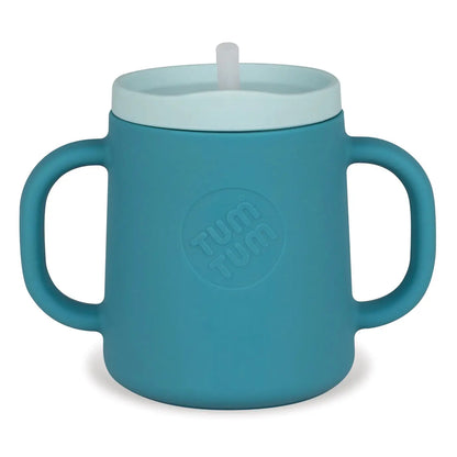 A teal blue silicone baby tumbler with integrated side handles, a matching lid, and a white straw spout. The brand name 'Tum Tum' is embossed in a circle on the front of the cup.