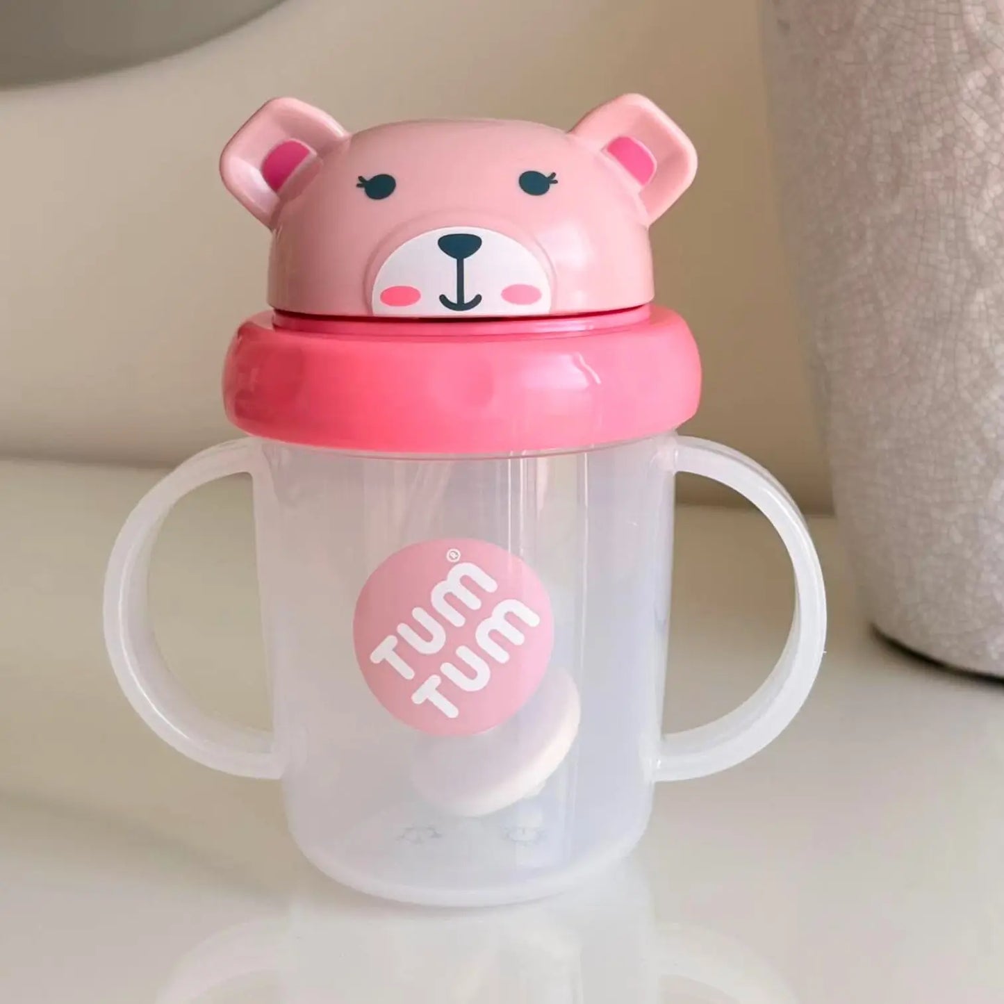 A clear plastic baby tumbler with white handles and a pink bear-shaped lid. The lid features a pink bear face with black eyes and nose, and a pink band around the neck.