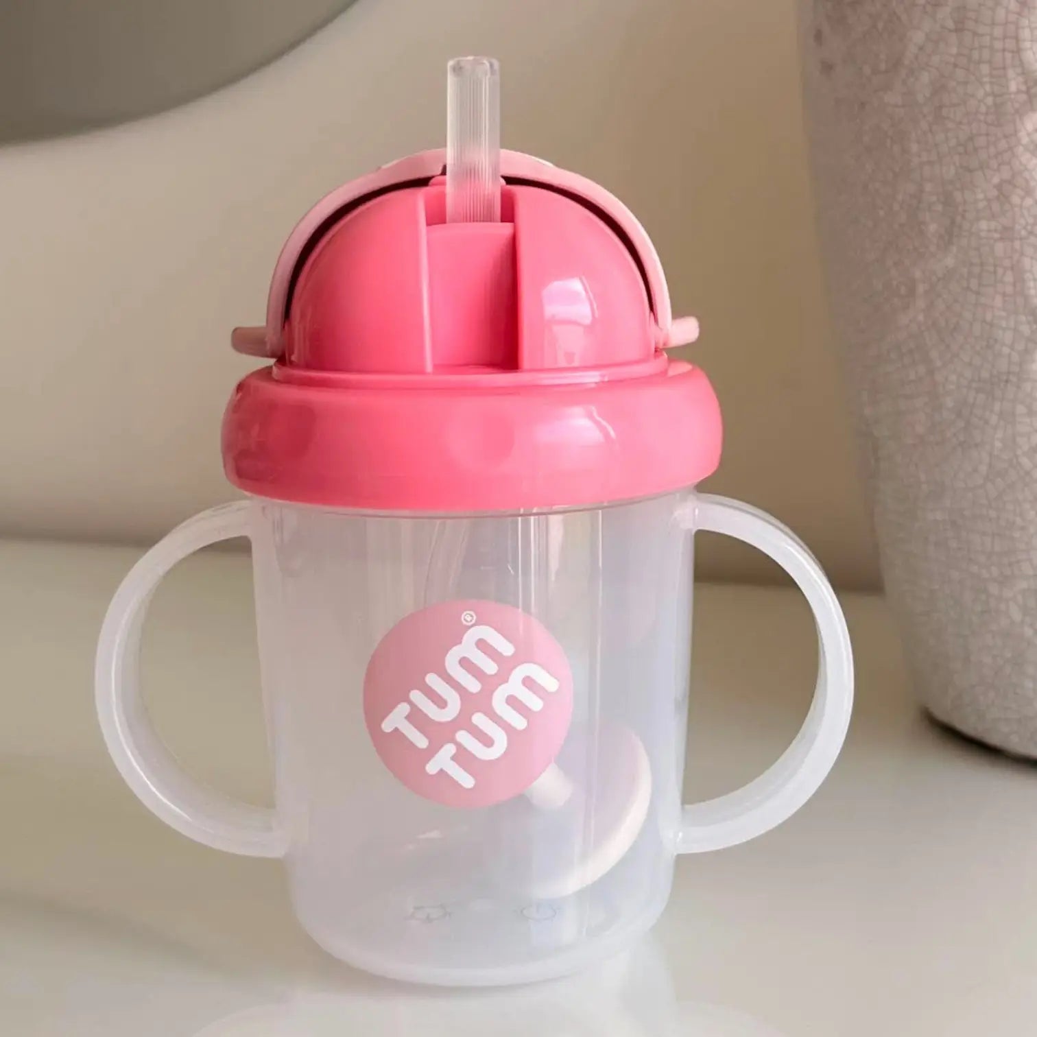 A pink and clear plastic baby tumbler cup with two white handles and a clear straw lid, featuring a pink circular 'TUM TUM' logo on the front, set against a soft beige background.