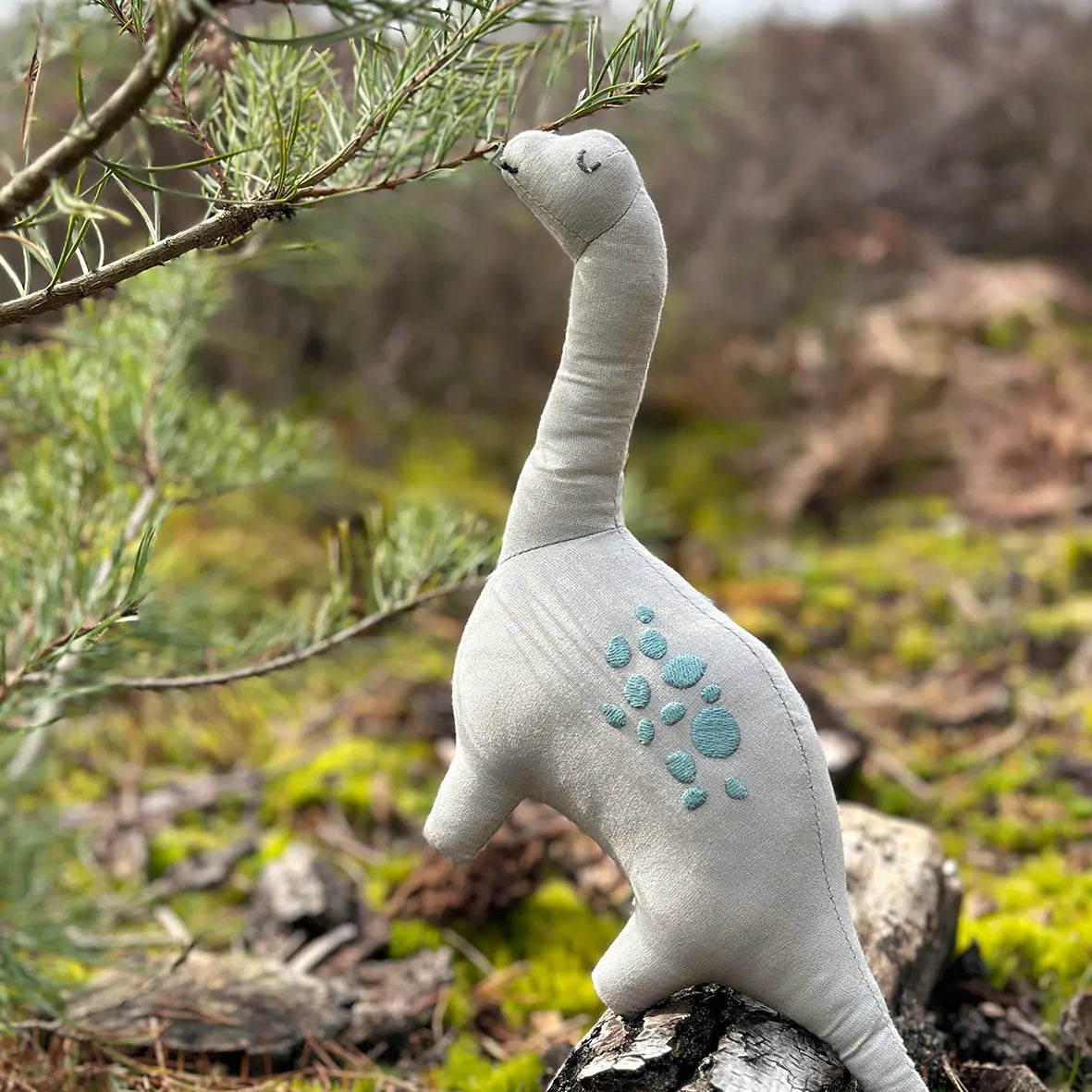 Plush dinosaur toy standing on a log in a natural setting with greenery.