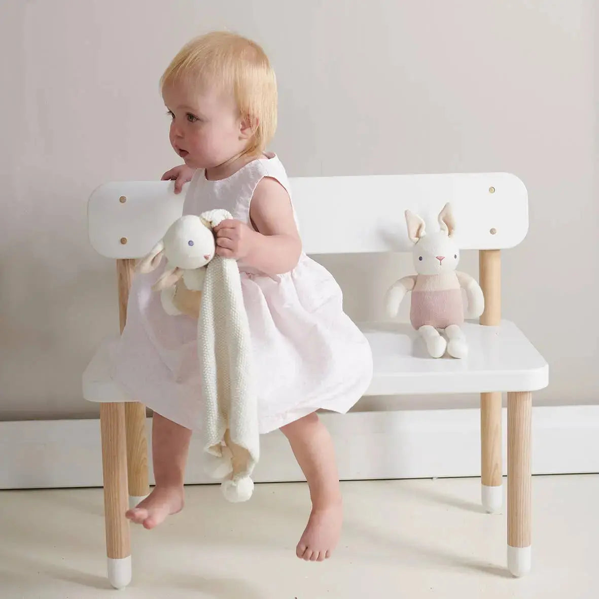 Child in a white dress sitting on a white chair with toys, against a plain background