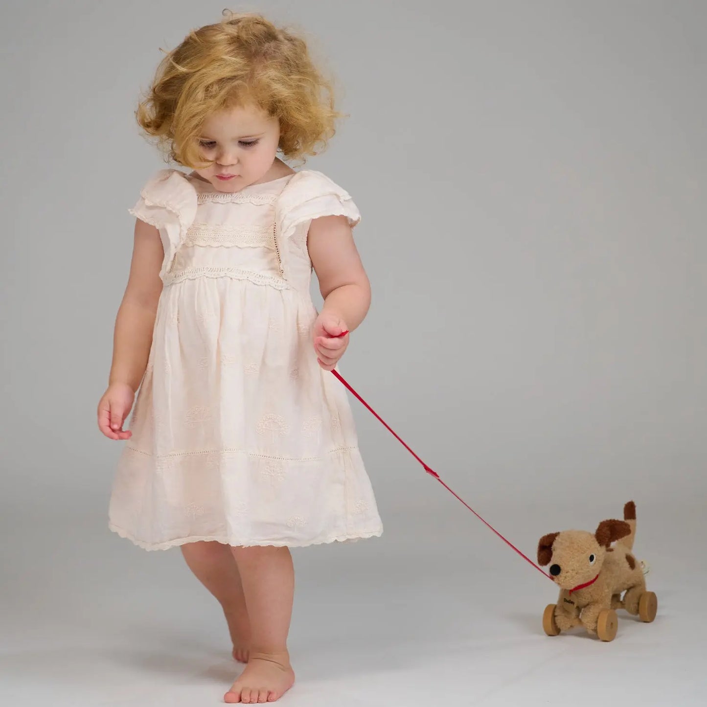 Child in a white dress holding a red leash attached to a small dog toy on a plain background