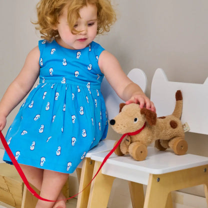 Child in a blue dress with white patterns holding a brown teddy bear on a red leash.