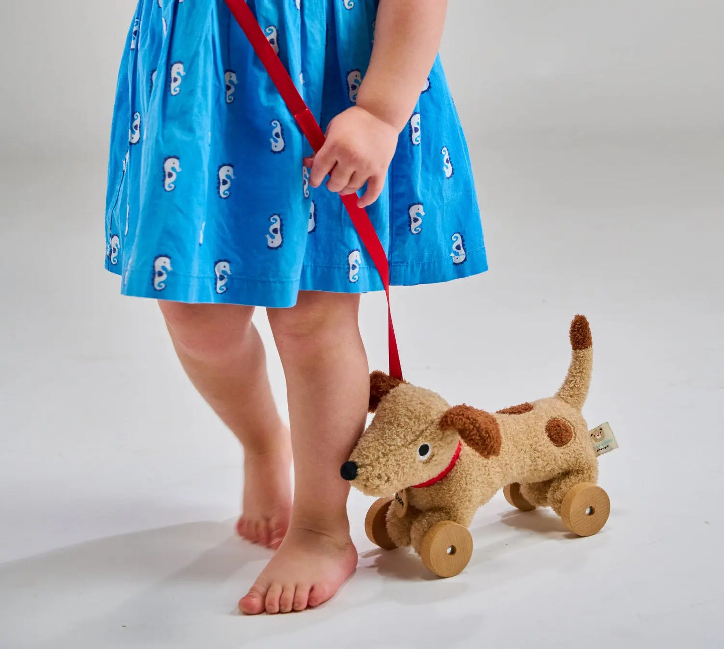 Child in a blue dress with white patterns holding a red leash attached to a brown toy dog on wheels.